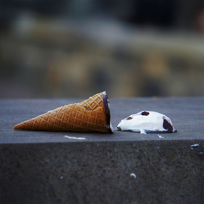 Eine Eiswaffel die auf einer Steinbank liegt, die Kugel liegt abgestürzt neben der Waffel