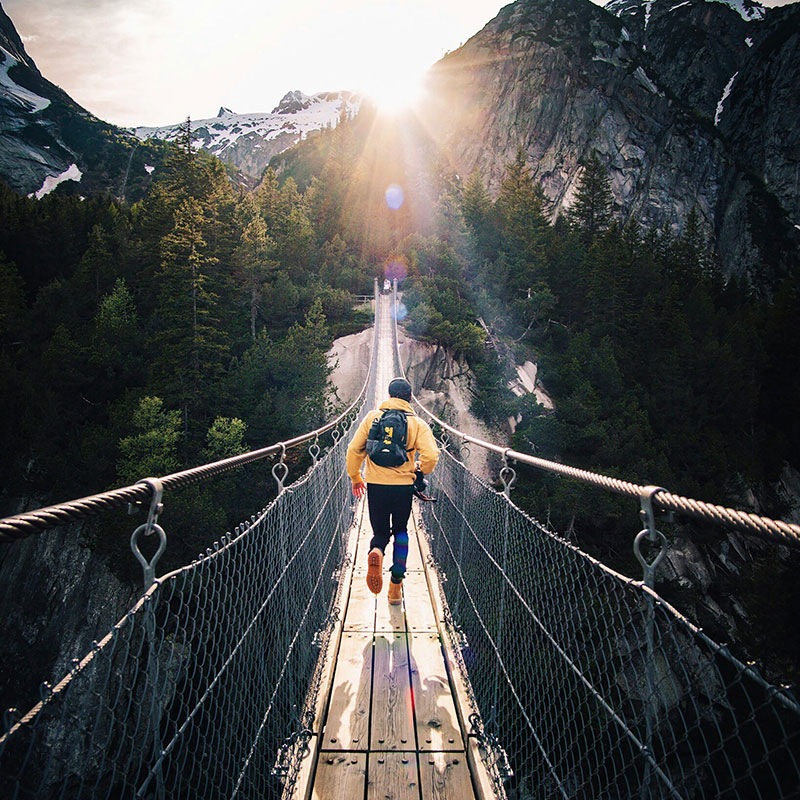 Ein Läufer mit Rucksack läuft auf einer Hängebrücke der Sonne entgegen